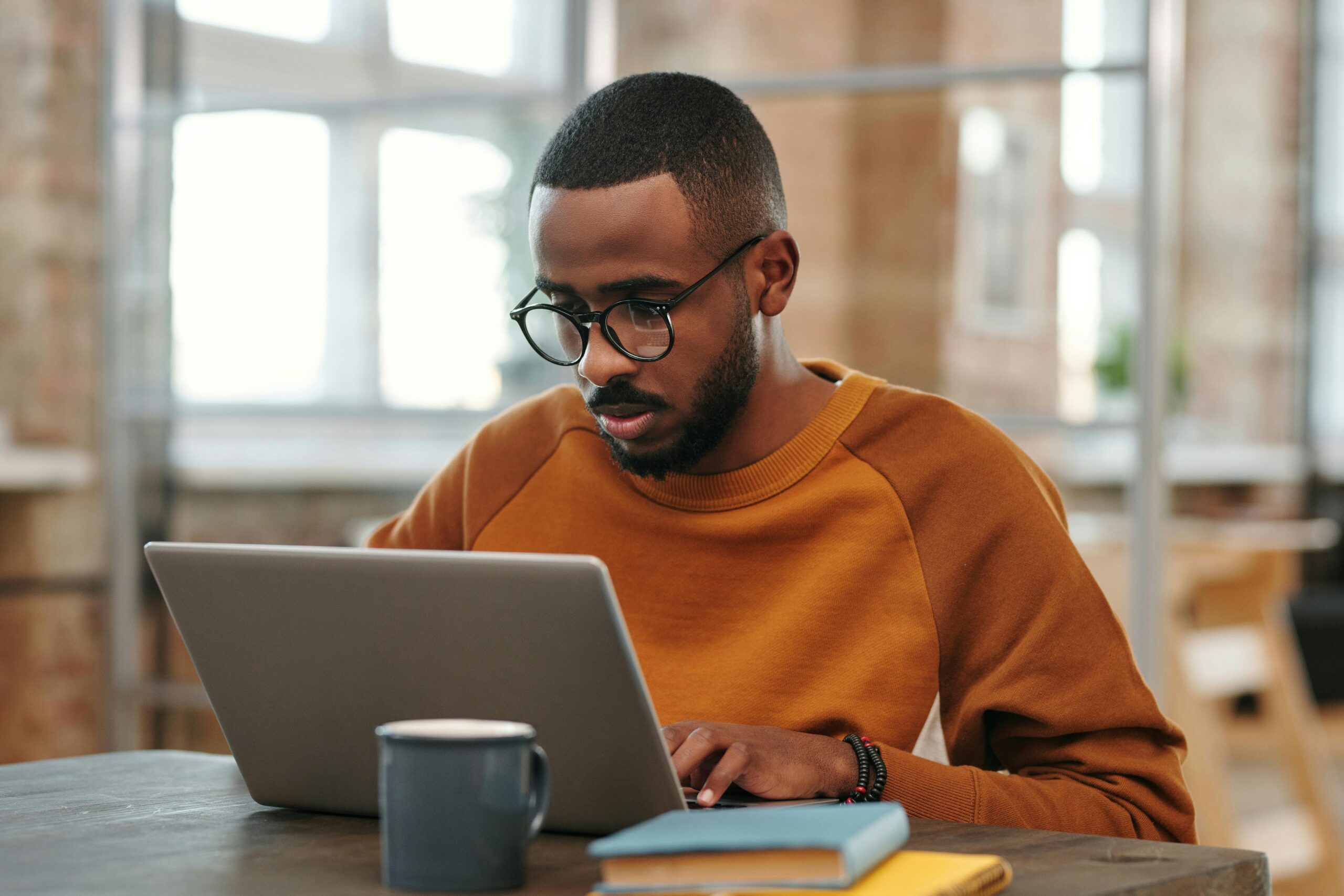 Young man with glasses works on laptop at a wooden table indoors. Warm, productive atmosphere.
